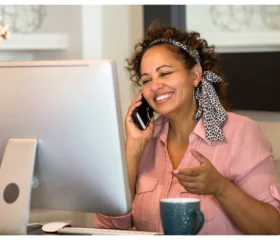 A woman wearing a pink blouse and patterned hair scarf smiles while talking on the phone at her desk, with a computer monitor and a blue mug in front of her.