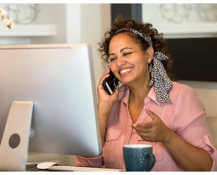 A woman wearing a pink blouse and patterned hair scarf smiles while talking on the phone at her desk, with a computer monitor and a blue mug in front of her.