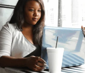 A woman with long hair sits at a table by a window, looking at a laptop. A white takeaway cup with a straw is on the striped table in front of her as she searches for a type 1 diabetes support group for women. Bright daylight comes through the window.