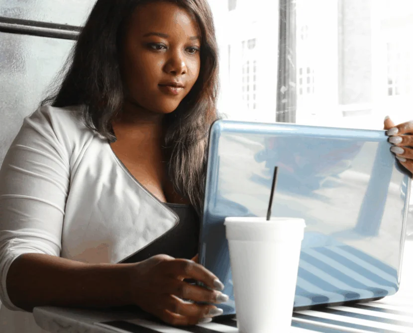 A woman with long hair sits at a table by a window, looking at a laptop. A white takeaway cup with a straw is on the striped table in front of her as she searches for a type 1 diabetes support group for women. Bright daylight comes through the window.