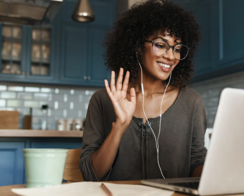 A woman with curly hair and glasses smiles and waves at her laptop during a video call, wearing earphones and sitting at a kitchen table with a mug, notepad, and pen.