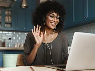 A woman with curly hair and glasses, wearing earphones, sits at a table and waves at her laptop screen, smiling. She appears to be on a video call in a modern kitchen.