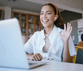A young woman wearing earphones and a red headband smiles and waves at her laptop during a video call, sitting indoors at a desk with a blurred library background.
