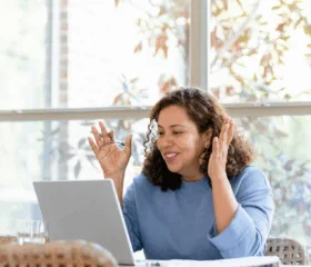 A woman with curly hair smiles and gestures while video chatting with her type 2 diabetes support group for women on a laptop at a table, with sunlight streaming through large windows and green plants visible outside.