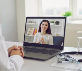 A doctor sits at a desk with a stethoscope and glass of water, having a video call with a smiling woman shown on the laptop screen.