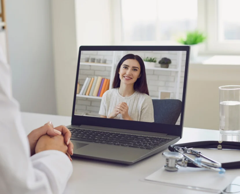 A doctor sits at a desk with a stethoscope and glass of water, having a video call with a smiling woman shown on the laptop screen.