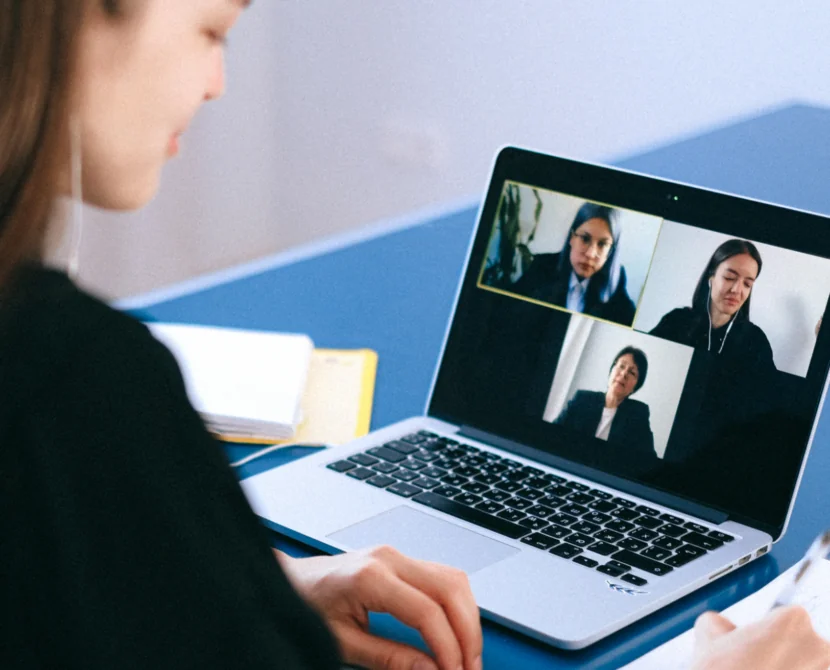A woman sits at a desk, taking notes while participating in a video conference call with three other people shown on her laptop screen.
