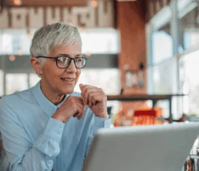 An older woman with short gray hair and glasses, wearing a light blue shirt, smiles while looking at a laptop in a bright, modern café, connecting with her type 1 diabetes support group for women.