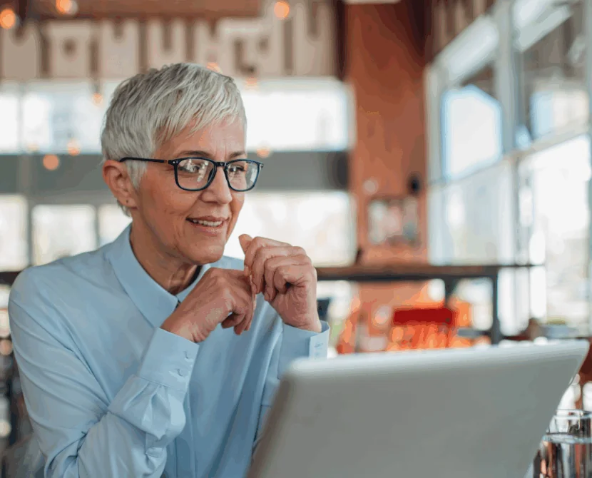 An older woman with short gray hair and glasses, wearing a light blue shirt, smiles while looking at a laptop in a bright, modern café, connecting with her type 1 diabetes support group for women.