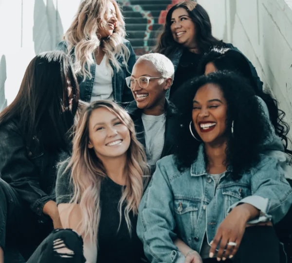 A group of six women, members of DiabetesSisters, sit together on outdoor steps, laughing and smiling. They wear casual denim and neutral clothing as sunlight brightens the scene, creating a joyful, friendly atmosphere.