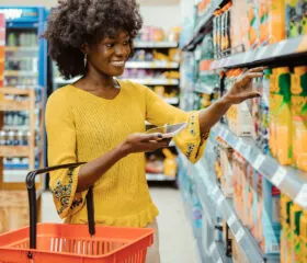 A woman in a yellow sweater holds a shopping basket and her phone while reaching for a carton of juice on a grocery store shelf. She is smiling and appears to be comparing products.