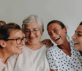 Four women of different ages stand close together, smiling and laughing warmly in front of a plain white background, reflecting the joy and togetherness found in a type 1 diabetes support group for women.