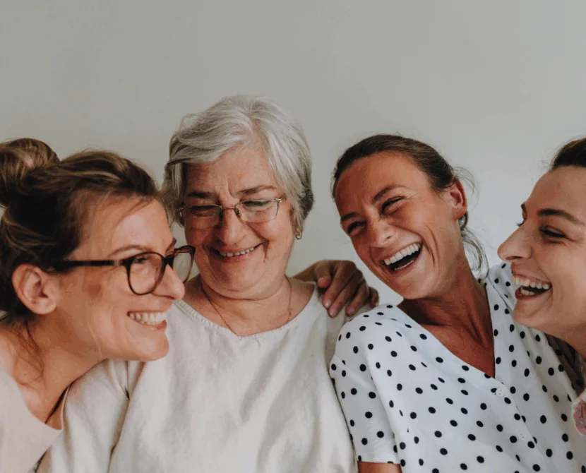 Four women of different ages stand close together, smiling and laughing warmly in front of a plain white background, reflecting the joy and togetherness found in a type 1 diabetes support group for women.