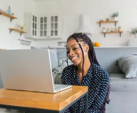 A woman with long braids sits on the floor, smiling at her laptop on a small table in a cozy, modern living room with shelves and light-colored decor in the background.