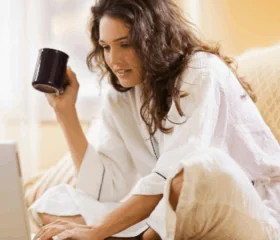 A woman in a white bathrobe sits on a couch holding a mug and looks at a laptop screen, appearing focused and relaxed in a cozy indoor setting.
