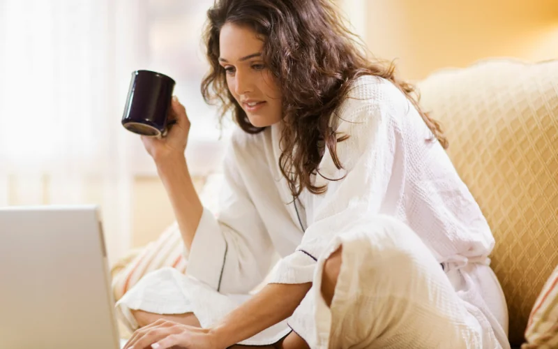 A woman in a white bathrobe sits on a couch holding a mug and looks at a laptop screen, appearing focused and relaxed in a cozy indoor setting.