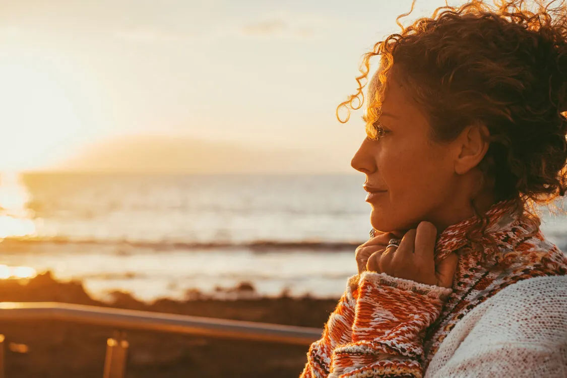 A woman with curly hair stands by the seaside at sunset, wearing a cozy sweater and looking thoughtfully into the distance, inspiring hope for women's health and diabetes foundation donations, as warm golden sunlight illuminates her face.