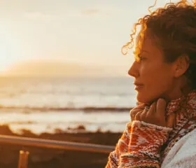 A woman with curly hair stands by the seaside at sunset, wearing a cozy sweater and looking thoughtfully into the distance, inspiring hope for women's health and diabetes foundation donations, as warm golden sunlight illuminates her face.