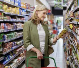 A woman in a green jacket holds a jar of pickles while shopping in a grocery store aisle, reading the label for her 7-day kidney health and diabetes guide. She has a red shopping basket and is surrounded by shelves filled with various food products.