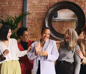 Five women stand together in front of a brick wall, smiling and laughing. They are wearing stylish, professional outfits and appear to be enjoying each others company in a friendly, relaxed setting.