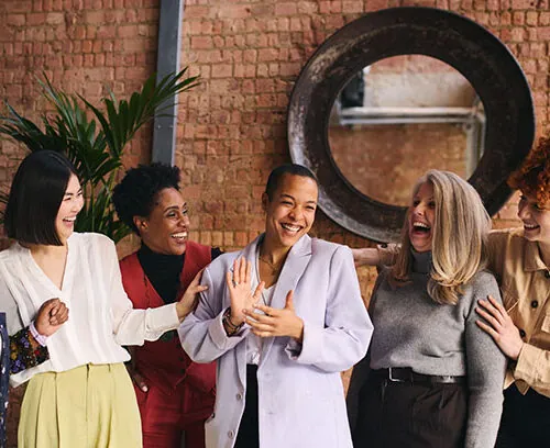 Five women stand together in front of a brick wall, smiling and laughing. They are wearing stylish, professional outfits and appear to be enjoying each others company in a friendly, relaxed setting.