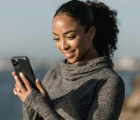 A woman wearing a gray long-sleeve top stands outdoors near water, smiling as she looks at her smartphone, possibly connecting with the DiabetesSisters community. The background is blurred, suggesting a sunny day by the coast.