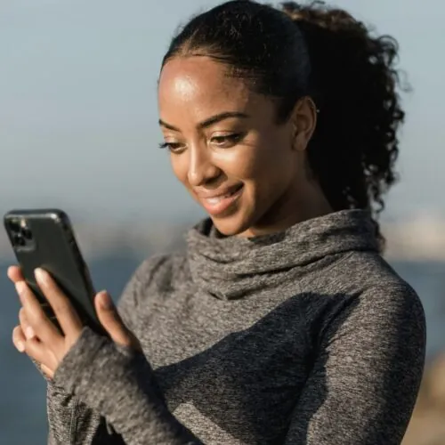 A woman wearing a gray long-sleeve top stands outdoors near water, smiling as she looks at her smartphone, possibly connecting with the DiabetesSisters community. The background is blurred, suggesting a sunny day by the coast.