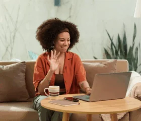A woman sits on a couch, smiling and waving at her laptop screen. A coffee mug, smartphone, and notebook are on the round wooden table in front of her. She appears to be on a video call in a bright, cozy setting.