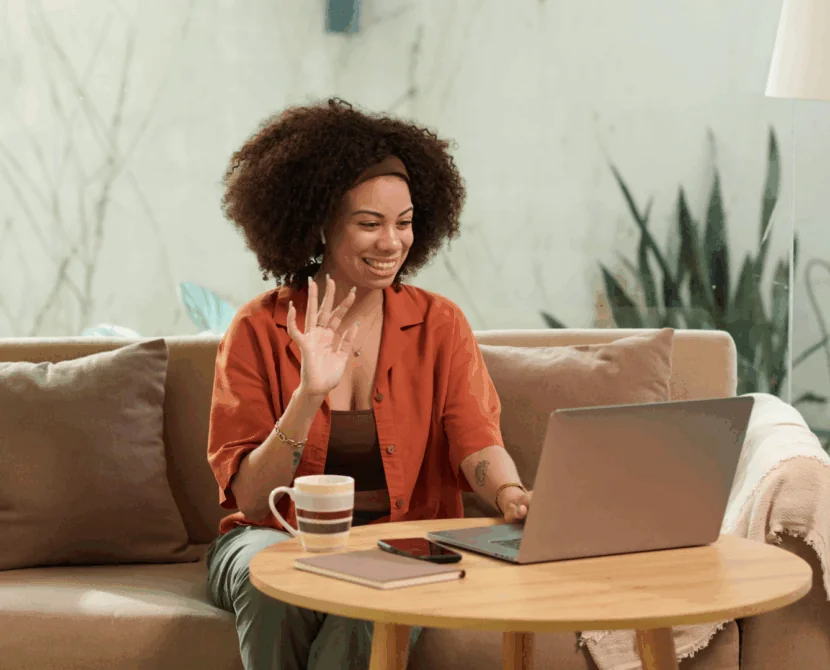 A woman sits on a couch, smiling and waving at her laptop screen. A coffee mug, smartphone, and notebook are on the round wooden table in front of her. She appears to be on a video call in a bright, cozy setting.
