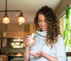 A woman with curly hair stands in a kitchen, smiling and looking at a pill in her hand while holding a glass of water. Warm light fixtures, green plants, and a diabetessisters support booklet add comfort to the inviting space.