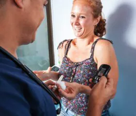A smiling woman in a floral top holds out her hand as a healthcare worker shows her a blood glucose meter, reflecting a positive health check guided by ADA Standards of Care for women with diabetes. The scene appears bright and friendly.