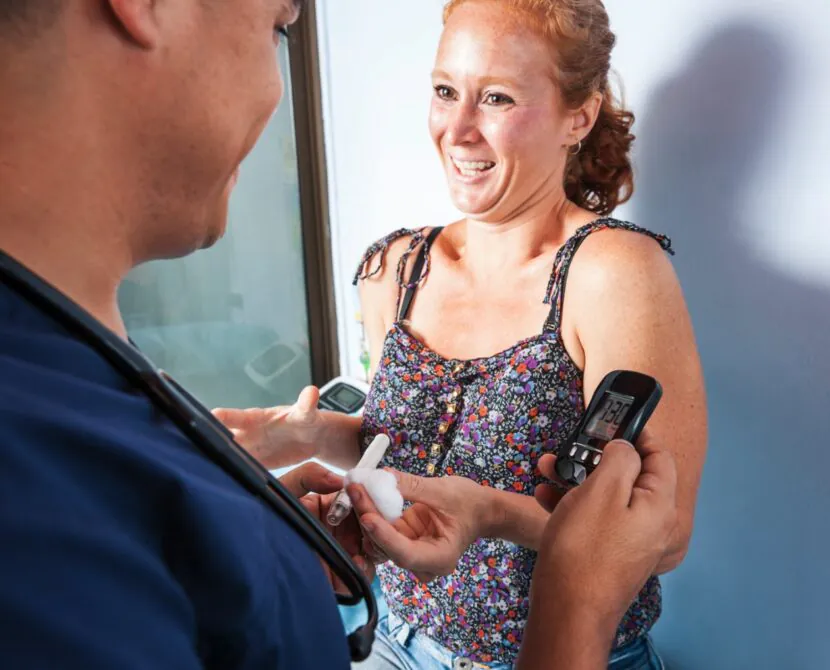 A smiling woman in a floral top holds out her hand as a healthcare worker shows her a blood glucose meter, reflecting a positive health check guided by ADA Standards of Care for women with diabetes. The scene appears bright and friendly.