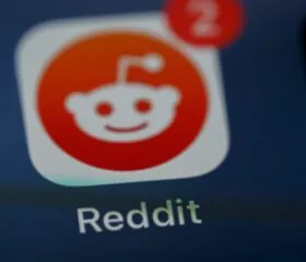 Close-up of the Reddit app icon on a device screen, with a red notification badge showing the number 2 in the upper right corner of the icon. The word Reddit appears below the icon.