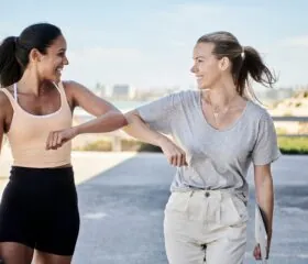 Two women smile and touch elbows in greeting while walking outdoors near a beach, wearing casual athletic clothes.
