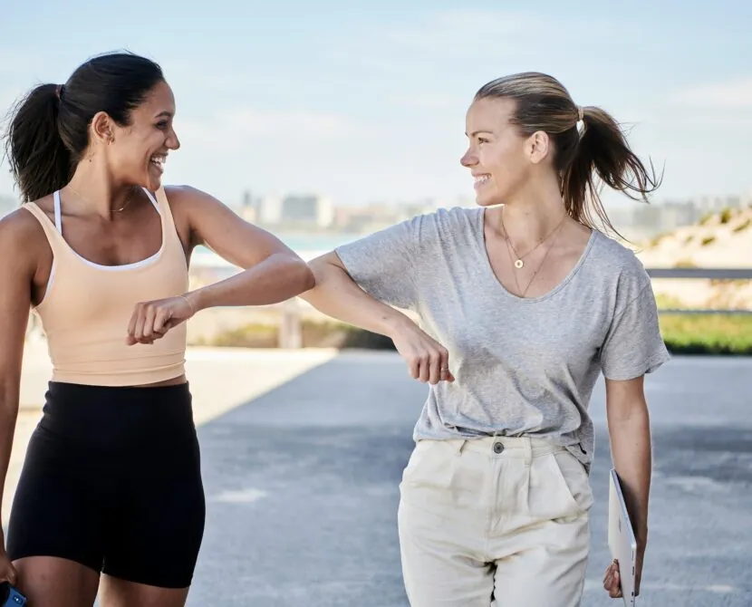 Two women smile and touch elbows in greeting while walking outdoors near a beach, wearing casual athletic clothes.