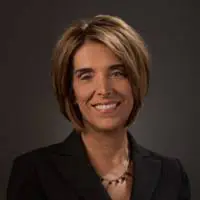 A woman with short, light brown hair wearing a black blazer and a beaded necklace, smiling in front of a dark, plain background.