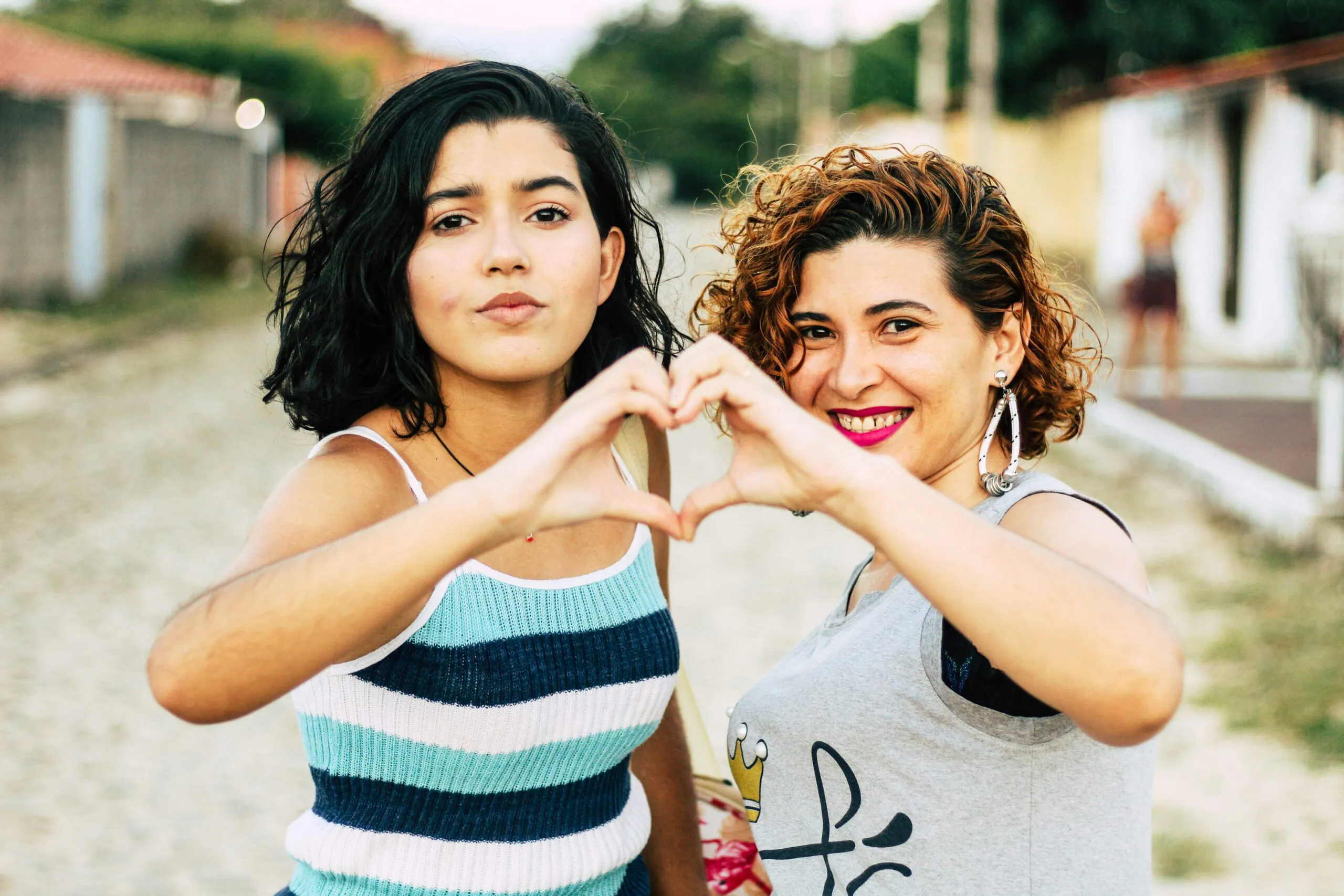 Two women stand close together outdoors, smiling and forming a heart shape with their hands toward the camera—celebrating la salud cardiovascular en mujeres con diabetes. One has short dark hair in a striped tank top; the other wears a light-colored top.