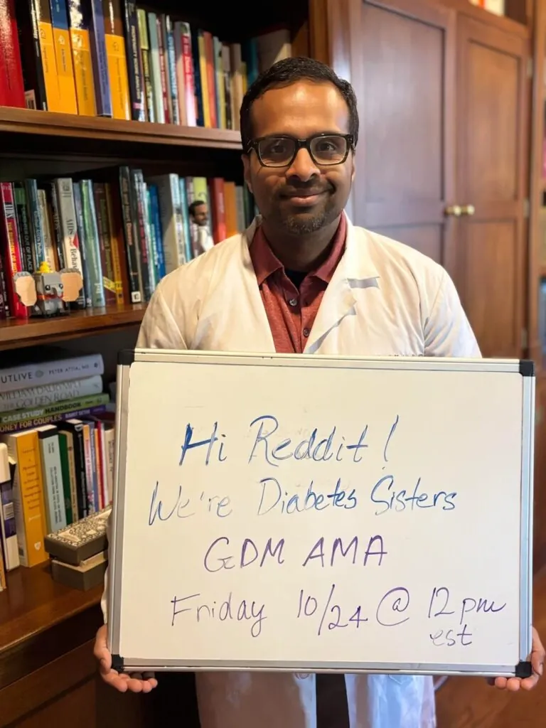 A man in a white coat stands in a library holding a sign that says, Hi Reddit! We’re Diabetes Sisters GDM AMA Friday 10/24 @ 12pm est. Join us to discuss your most asked questions about gestational diabetes.