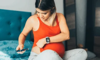 A pregnant woman in an orange tank top sits on a bed, focused as she checks her blood sugar with a glucometer—part of managing gestational diabetes. She wears a smartwatch and presses the device to her abdomen, monitoring her health closely.