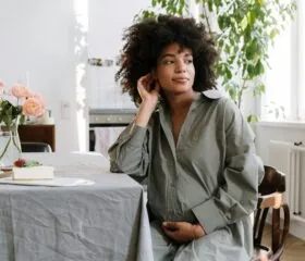 A woman with curly hair sits at a dining table, touching her ear and resting her hand on her stomach. She wears a loose gray shirt and looks thoughtfully to the side. There is a slice of cake and flowers on the table.
