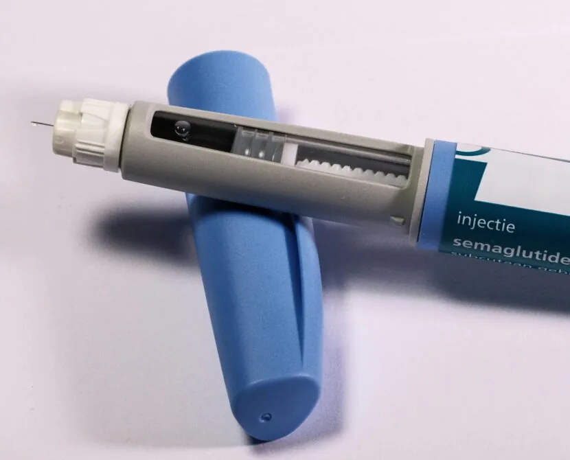 A close-up of a semaglutide injection pen with a visible needle, placed on a white surface next to its blue protective cap.