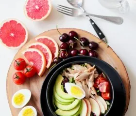 A colorful meal with sliced avocado, boiled egg, tomatoes, shredded chicken, cherries, grapefruit, and a bowl on a wooden board, next to cutlery and a glass on a white surface.