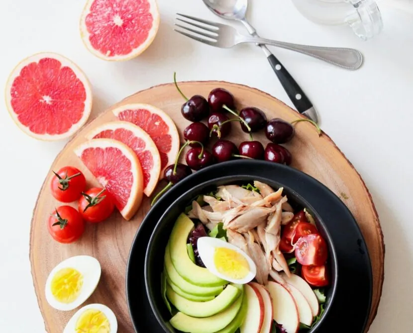 A colorful meal with sliced avocado, boiled egg, tomatoes, shredded chicken, cherries, grapefruit, and a bowl on a wooden board, next to cutlery and a glass on a white surface.