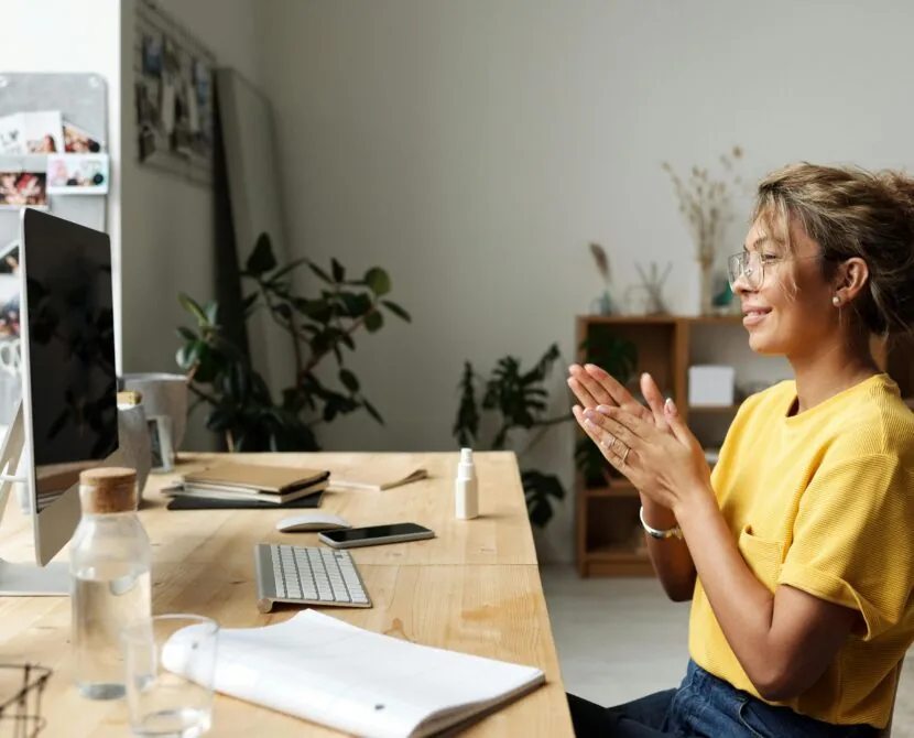 A woman wearing glasses and a yellow shirt sits at a desk, smiling and clapping while looking at a computer screen—perhaps tuning into a DiabetesSisters virtual event—in a bright, modern home office with plants and shelves in the background.