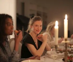Women sharing a cozy candlelit dinner, with one woman smiling warmly toward the camera while others enjoy the meal.