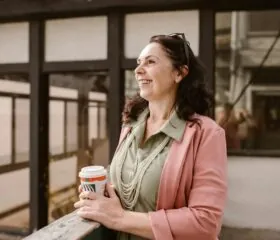 A smiling woman in a pink blazer and green shirt holds a takeout coffee cup and stands outdoors, leaning on a wooden rail, with sunglasses on her head and buildings in the background.