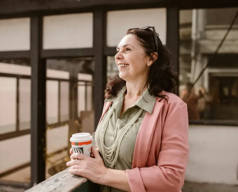 A smiling woman in a pink blazer and green shirt holds a takeout coffee cup and stands outdoors, leaning on a wooden rail, with sunglasses on her head and buildings in the background.