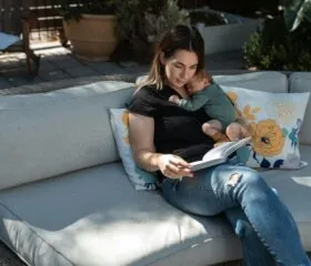A woman sits on an outdoor couch reading a book while holding a sleeping baby on her chest. The scene is calm, with sunlight and decorative floral pillows around them.