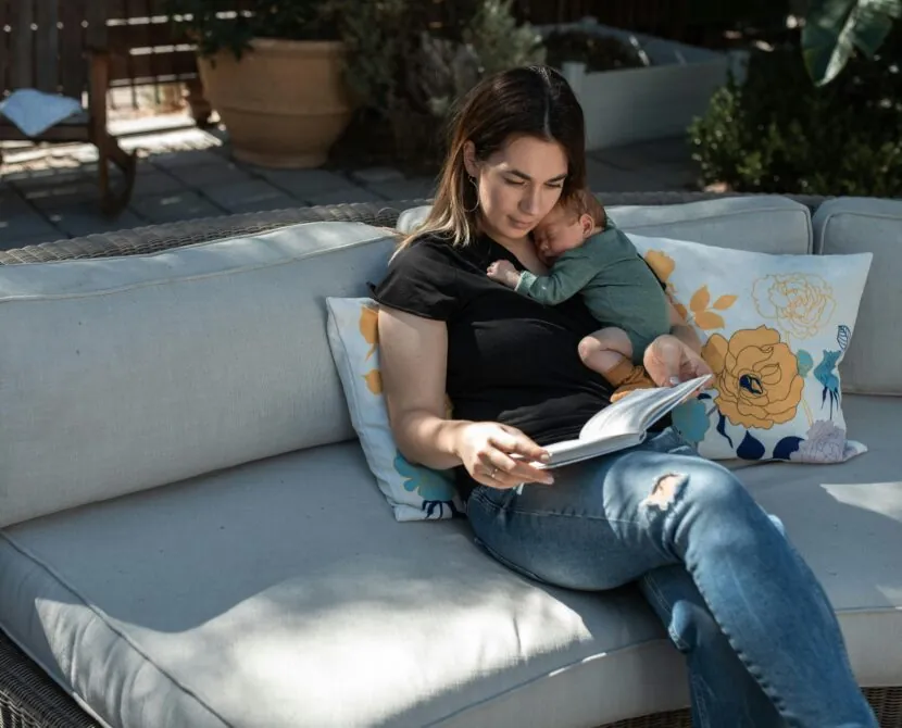 A woman sits on an outdoor couch reading a book while holding a sleeping baby on her chest. The scene is calm, with sunlight and decorative floral pillows around them.