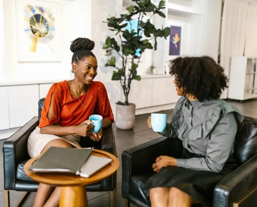 Two women sit in armchairs, smiling and talking while holding blue mugs. A small round table with a laptop and notebook is between them, reflecting the supportive community of Diabetessisters in a modern, brightly lit office or lounge area.